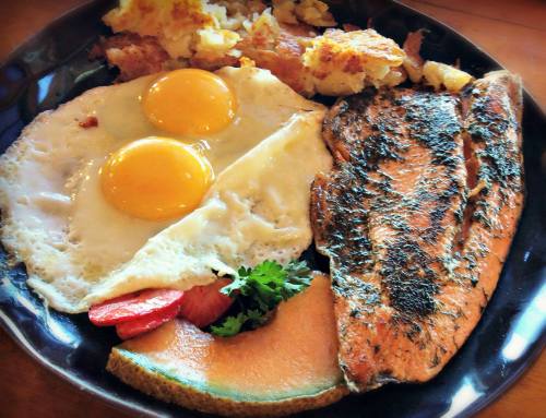 A plate of breakfast featuring two sunny-side-up eggs, grilled fish, hash browns, strawberries, and cantaloupe.
