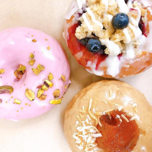 Three colorful donuts: one pink with pistachios, one with blueberries and icing, and one with coconut and apricot filling.