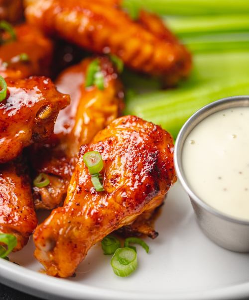 A plate of glazed chicken wings garnished with green onions, served with celery sticks and a small cup of dipping sauce.