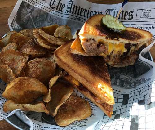 A grilled sandwich with melted cheese and pickles, served with a side of crispy potato chips on a newspaper-lined tray.