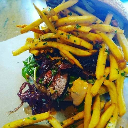 A hearty burger topped with crispy fries, greens, and sauce, served on a wooden table.