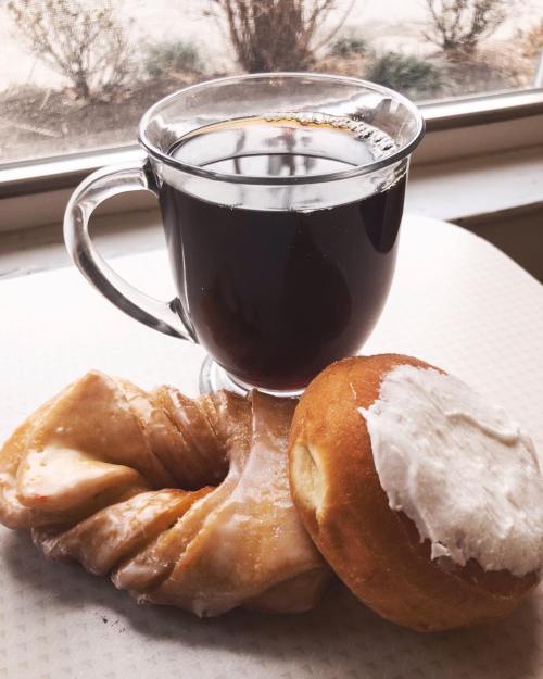 A cup of black coffee next to a glazed pastry and a frosted donut on a table by a window.