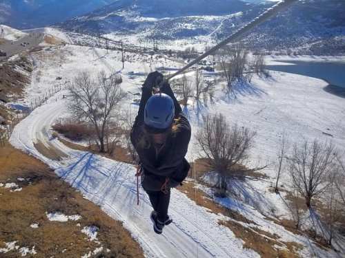 A person zip-lining over a snowy landscape with mountains and a winding road below.