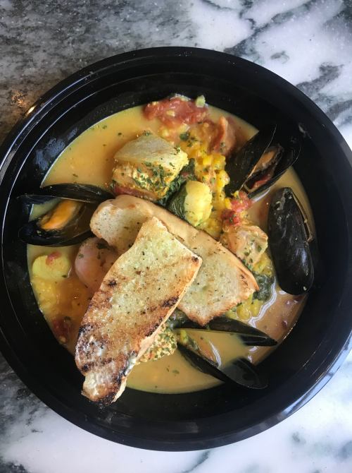 A bowl of seafood stew with mussels, vegetables, and toasted bread on top, served on a marble surface.