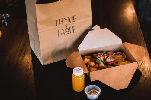 A takeout box with food, a small bottle of juice, and a paper bag labeled "THYME x TABLE" on a dark wooden table.