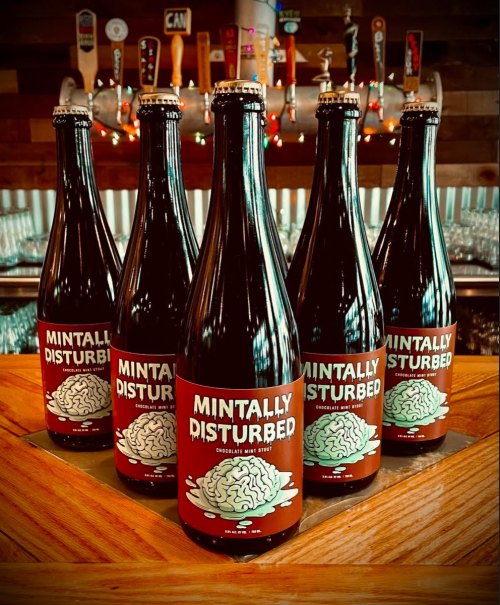 Five bottles of "Mintally Disturbed" chocolate mint stout on a wooden bar, with colorful beer taps in the background.