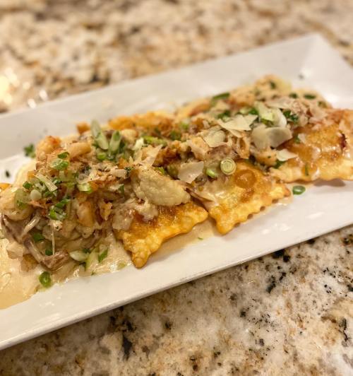 A plate of ravioli topped with shredded chicken, green onions, and crispy bits, served on a marble countertop.