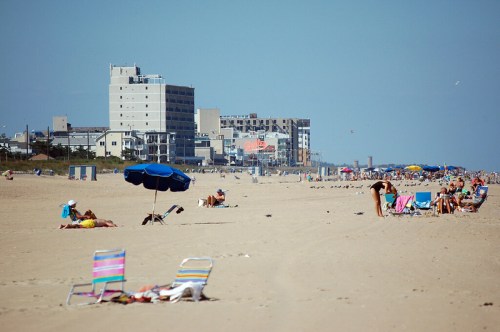 A sandy beach with colorful umbrellas and chairs, people relaxing, and buildings in the background under a clear blue sky.