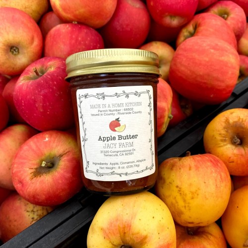 A jar of apple butter sits among fresh apples, labeled "Jacy Farm" with ingredients listed on the front.