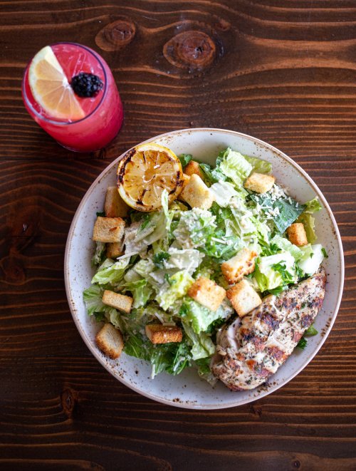 A bowl of Caesar salad with grilled chicken, croutons, and a lemon slice, alongside a pink drink with a lemon wedge.