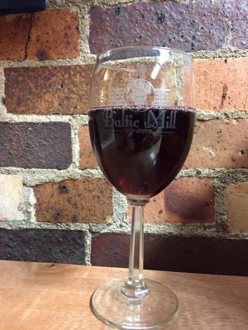A glass of red wine sits on a wooden table in front of a brick wall, featuring the Baltic Mill Winery logo.