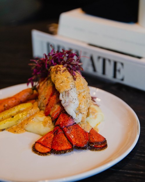 A gourmet dish featuring lobster tail on mashed potatoes, garnished with purple microgreens and colorful roasted vegetables.