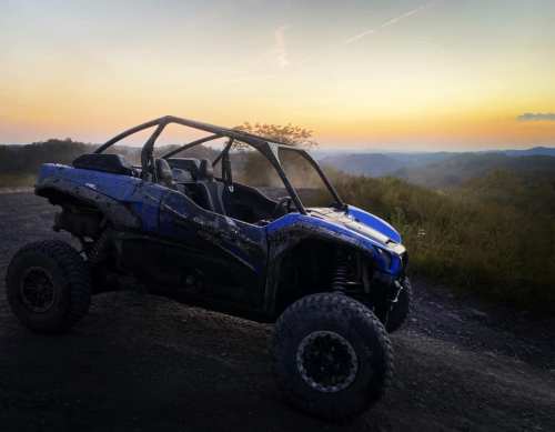 A blue off-road vehicle parked on a dirt path with a sunset and rolling hills in the background.