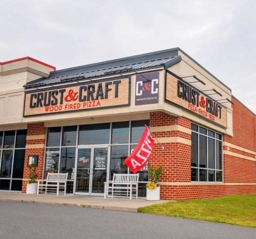 Exterior of Crust & Craft, a wood-fired pizza restaurant, featuring a red flag and outdoor seating.