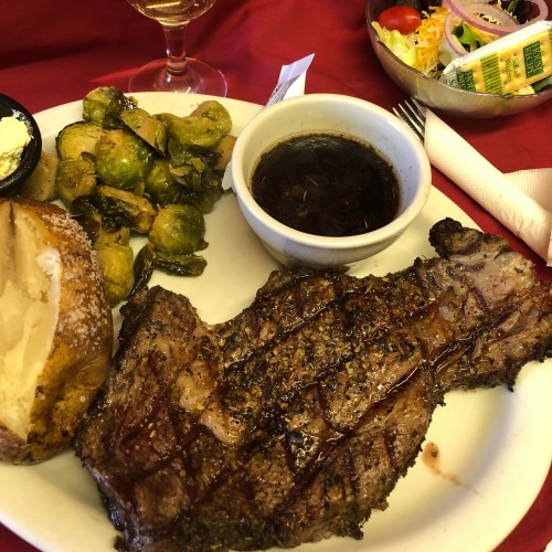 A plate featuring a grilled steak, Brussels sprouts, a baked potato, and a side of dipping sauce, with a salad in the background.