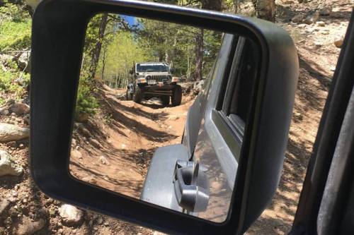 A rugged Jeep is seen through the side mirror of another vehicle on a rocky, forested trail.