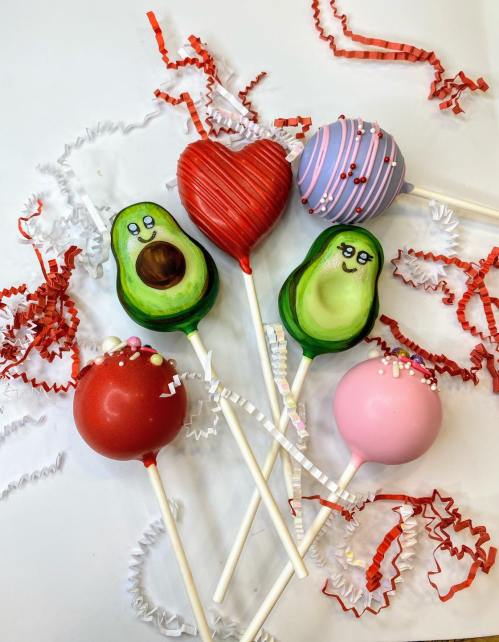 Colorful cake pops shaped like avocados, hearts, and round designs, surrounded by festive red and white crinkle paper.