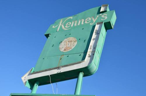 A weathered green sign for "Kenney's" against a clear blue sky, showing signs of age and wear.