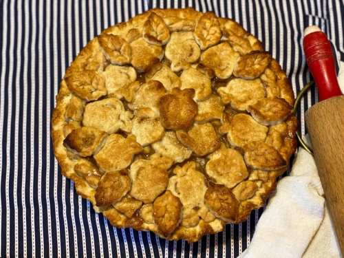 A golden-brown pie with decorative leaf-shaped crust on a striped cloth, accompanied by a rolling pin and towel.