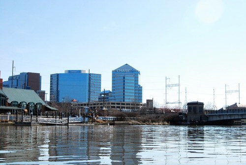 City skyline featuring modern buildings and a river in the foreground on a clear day.