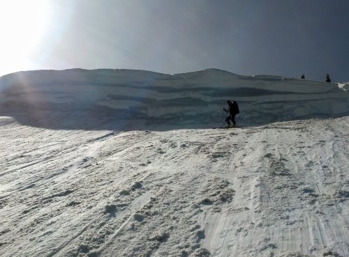 A person climbs a snowy slope with a backpack, surrounded by a bright, sunny sky and snow-covered terrain.