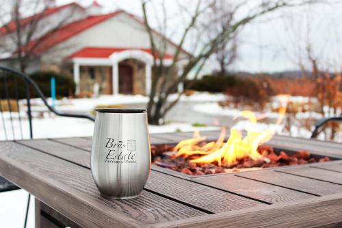 A stainless steel mug sits on a table by a fire pit, with a house and snowy landscape in the background.