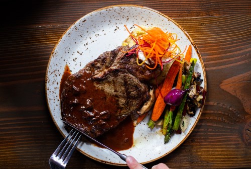 A plate of steak with rich sauce, garnished with colorful vegetables and carrot curls, served on a rustic wooden table.