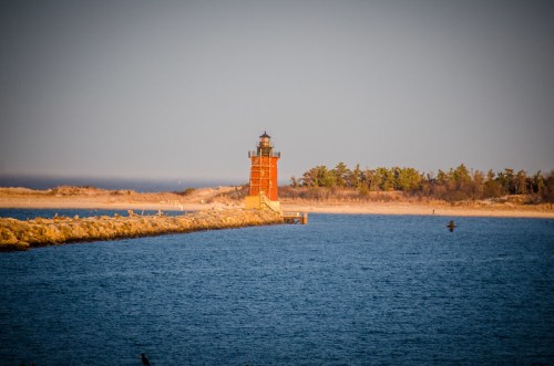 A red lighthouse stands on a rocky pier, surrounded by calm blue water and a sandy shore in the background.