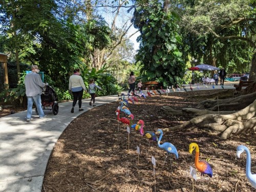 A winding path in a lush park lined with colorful flamingo decorations, with people walking and enjoying the scenery.