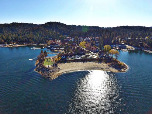 Aerial view of a scenic lakeside area with a sandy beach, surrounded by trees and buildings on a sunny day.