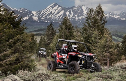 Two riders in a red ATV navigate a mountainous trail, surrounded by trees and snow-capped peaks in the background.