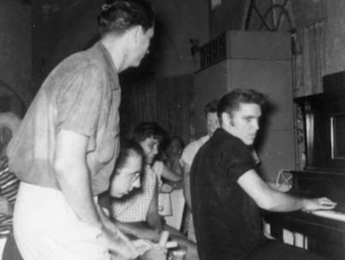 Elvis Presley plays piano surrounded by a group of people in a vintage setting, capturing a moment of musical collaboration.