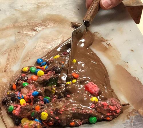 A hand holding a spatula mixes colorful candy into melted chocolate on a marble surface.