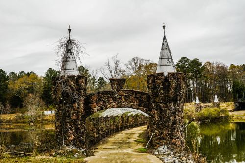 A stone bridge with towers leads over a path by a lake, surrounded by trees under a cloudy sky.