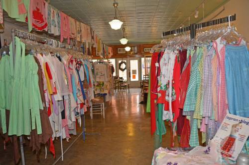 A colorful display of vintage clothing hangs on racks in a shop, with tables and chairs visible in the background.