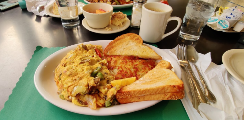 A plate of scrambled eggs with vegetables, hash browns, and toast, with drinks and other dishes in the background.