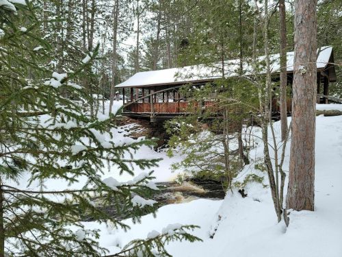 A wooden bridge over a snowy landscape, surrounded by tall trees and a flowing stream.