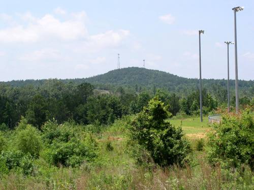A green landscape with a hill in the background, featuring communication towers and tall light poles in the foreground.