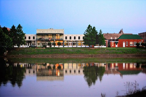 A serene riverside view of buildings reflecting on the water at dusk, with trees lining the shore.