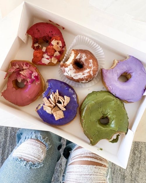 A box of six colorful donuts, featuring pink, purple, and green icing, with various toppings and one partially eaten.