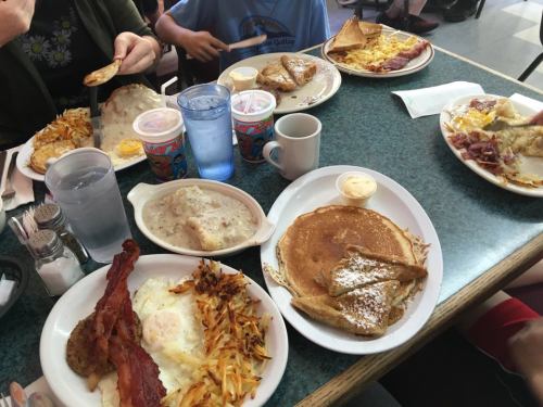 A table filled with breakfast plates, including pancakes, eggs, bacon, hash browns, and drinks in a diner setting.