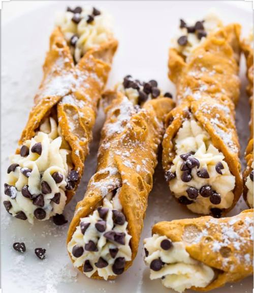 Five crispy cannoli filled with sweet cream and mini chocolate chips, dusted with powdered sugar on a white plate.