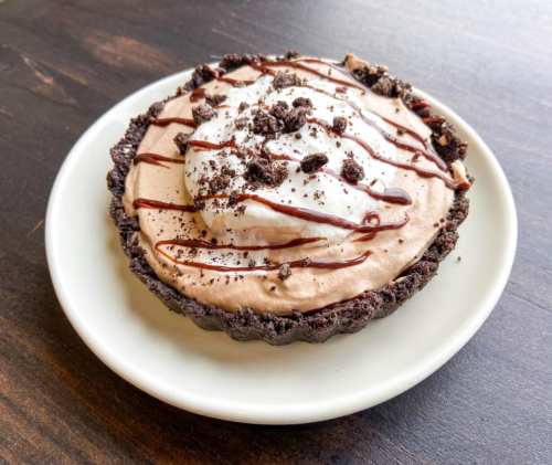 A chocolate cream pie in an Oreo crust, topped with whipped cream and chocolate drizzle, served on a white plate.
