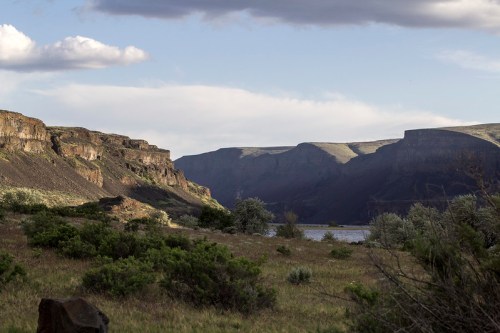 A scenic landscape featuring cliffs, a river, and lush greenery under a partly cloudy sky.