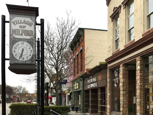 Sign for "Village of Milford" with shops and trees lining a street on a cloudy day.