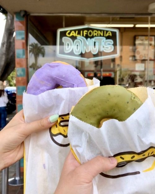 Two hands hold colorful donuts—one purple and one green—wrapped in paper, with a donut shop sign in the background.