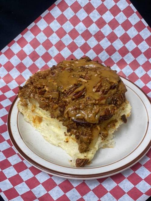 A slice of dessert topped with a rich, nutty caramel sauce and pecans, served on a plate with a checkered background.