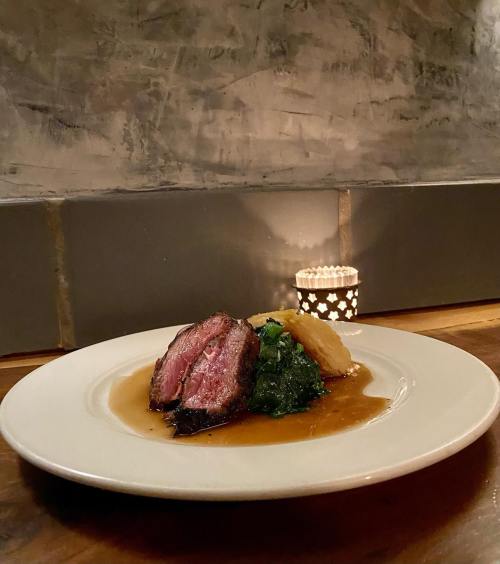 A plate of sliced beef with greens and potatoes, served with sauce, next to a lit candle on a rustic wooden table.