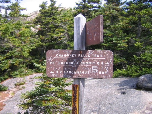 Trail signpost in a forest, indicating Champney Falls Trail, Mt. Chocorua Summit, and Kancamagus Highway directions.