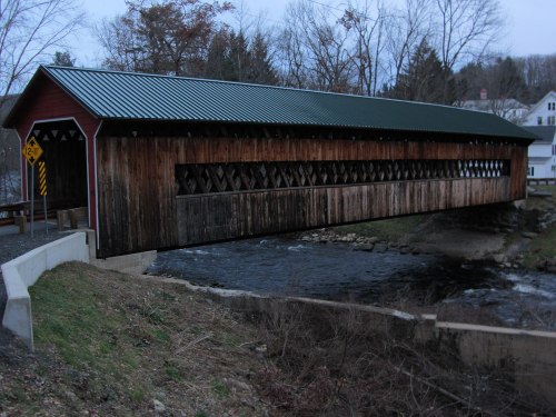 A wooden covered bridge spans a river, surrounded by trees and a small town in the background, during twilight.
